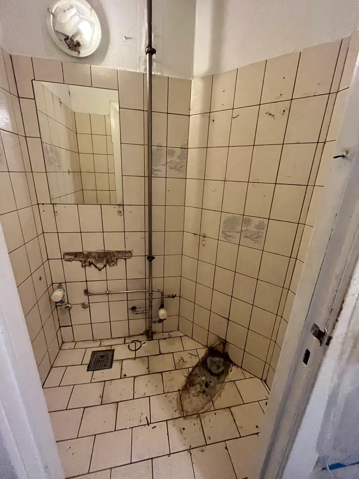Slidt bruserum med gamle cremefarvede fliser, lodret stålrør fra gulv til loft, gulvafløb i hjørnet og opbrudt hul i det gamle gulv